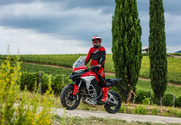 Un motociclista in tuta rossa sfreccia tra i cipressi e i vigneti del Val d'Orcia in moto.