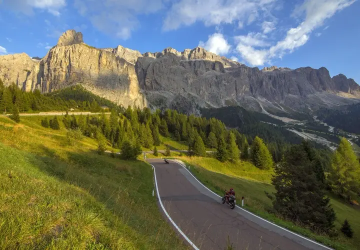 Strada tortuosa tra prati verdi e boschi di conifere, con imponenti vette dolomitiche sullo sfondo. Due motociclisti godono del panorama mozzafiato, un'avventura indimenticabile tra le Dolomiti in moto.