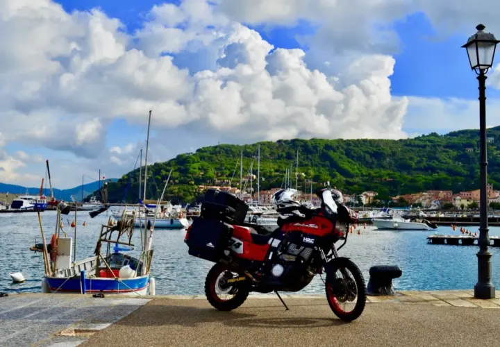 Moto pronta per l'avventura: Isola d'Elba in moto 3 giorni. Porto con barche e collina verde sullo sfondo.