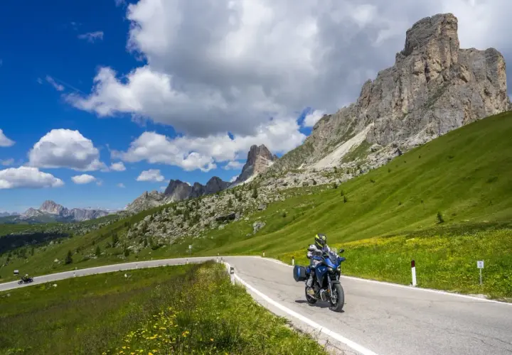 Motociclista percorre una strada panoramica tra i laghi del Veneto, con montagne imponenti e cielo azzurro.