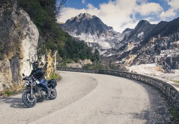 Motociclista su strada panoramica toscana, con montagne di marmo sullo sfondo.