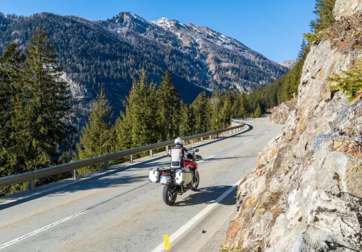 Un motociclista percorre una strada tortuosa in Valtellina, circondato da imponenti montagne e fitti boschi di conifere.