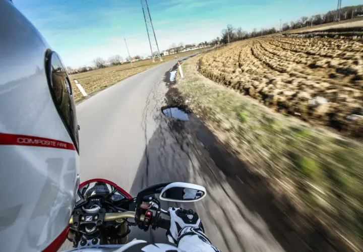 In sella a una moto, con il casco bianco e rosso, si percorre una strada di campagna. Il campo arato a lato e il cielo azzurro evocano il linguaggio motociclisti italiano.