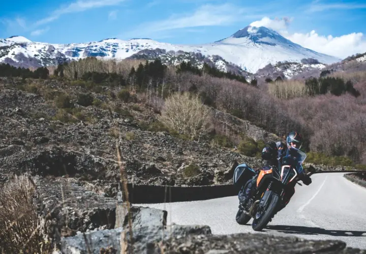 Motociclista su strada mareneve, con vulcano innevato sullo sfondo.