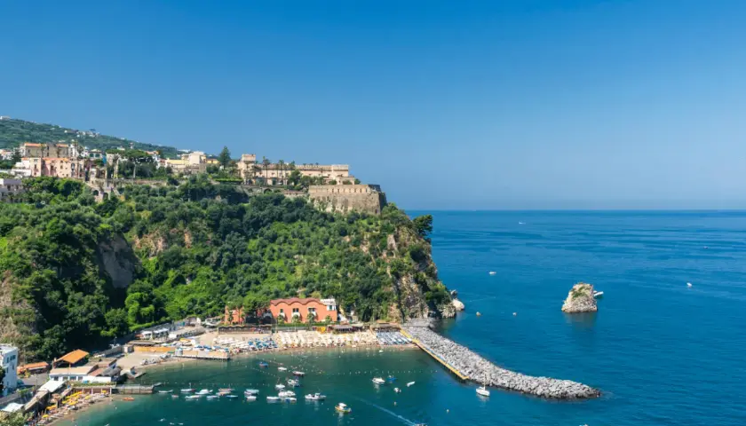 Vico Equense cosa vedere: una baia con spiaggia attrezzata, un molo di rocce e un borgo arroccato sulla scogliera.