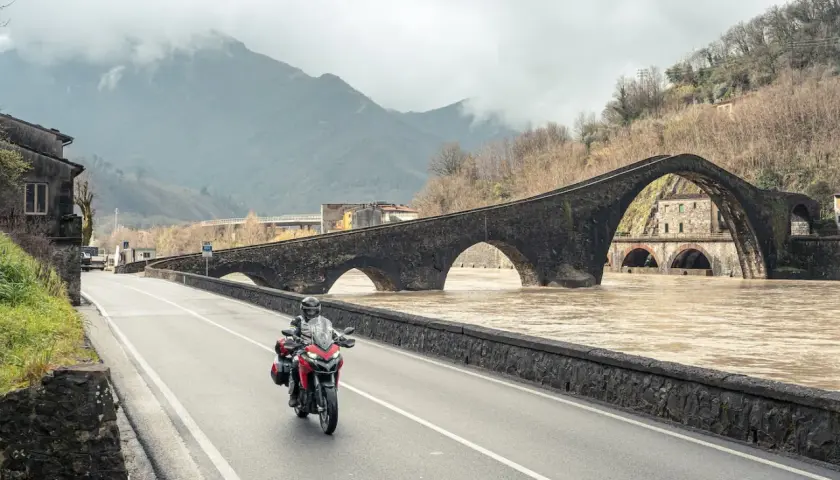 Motociclista attraversa il Ponte del Diavolo, un viaggio tra Lucca e Siena, con il fiume in piena sotto le arcate.