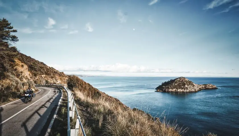 Un motociclista percorre una strada panoramica sull'Argentario, con il mare blu e un'isola rocciosa sullo sfondo.