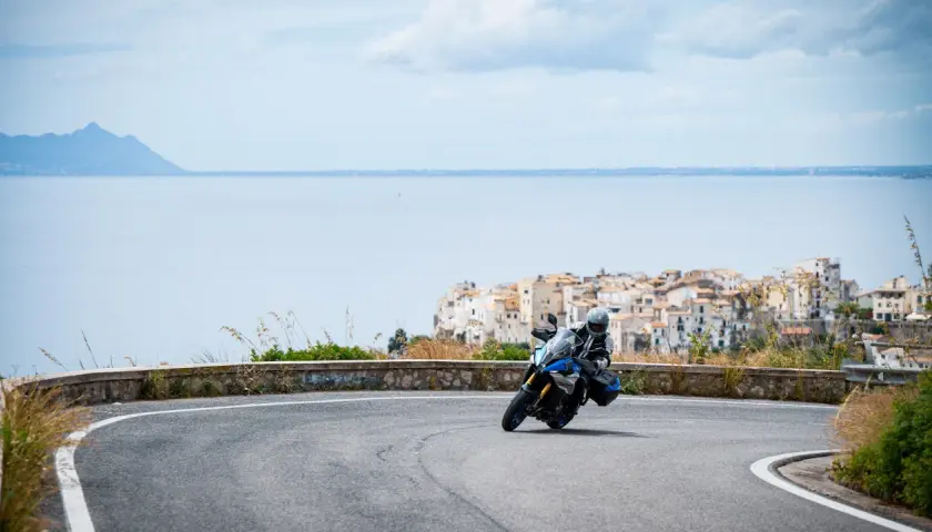 Motociclista in curva su strada panoramica, con il mare e il borgo di Sperlonga sullo sfondo. Un viaggio da Terracina a Sperlonga.