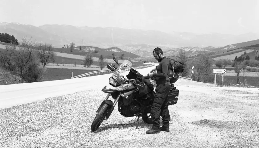 Motociclista con zaino e attrezzatura da viaggio si ferma accanto alla sua moto, la sella ciampigotto pronta per la prossima avventura. Paesaggio collinare sullo sfondo.