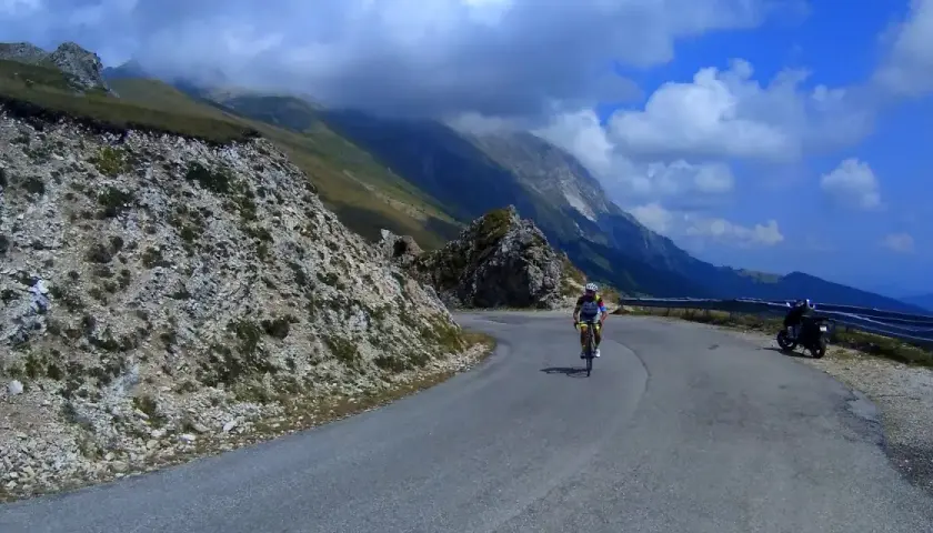 Ciclista in salita sulla strada panoramica della Forca di Presta, con montagne sullo sfondo e una moto parcheggiata.