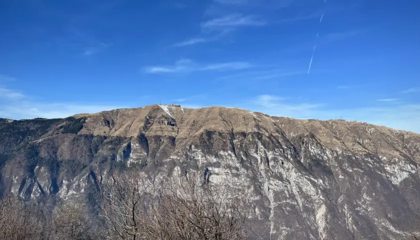 Vetta del monte Pizzoc con neve residua, pendii bruni e rocce bianche sotto un cielo azzurro con scie di aerei.