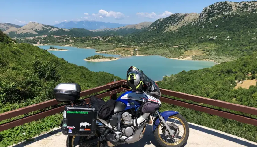Moto pronta per esplorare gli Appennini, con un lago turchese e montagne verdi sullo sfondo. #AppenniniInMoto