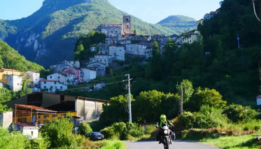 Un motociclista esplora la Lunigiana, con un borgo arroccato su una collina verdeggiante sullo sfondo.