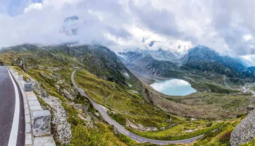 Strada tortuosa sul passo della Sambuca, con lago alpino azzurro e montagne innevate sullo sfondo.