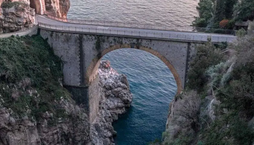 Ponte ad arco in pietra che attraversa un fiordo, con il mare blu sottostante. Un'icona del furore costiera amalfitana, incastonata tra rocce e vegetazione.