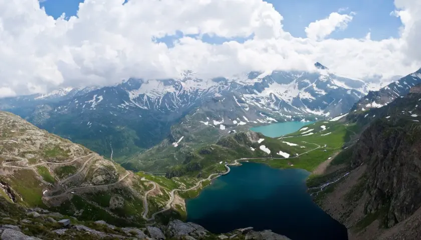 Strada tortuosa sul passo Nivolet, con laghi alpini e montagne innevate sotto un cielo nuvoloso.