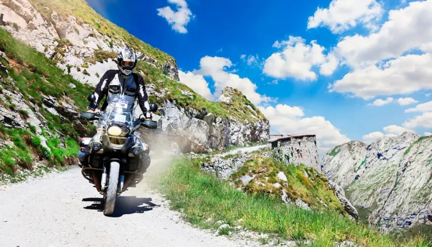 Motociclista affronta un ripido tornante della boaria su una strada sterrata in montagna, con un cielo azzurro e nuvole bianche.