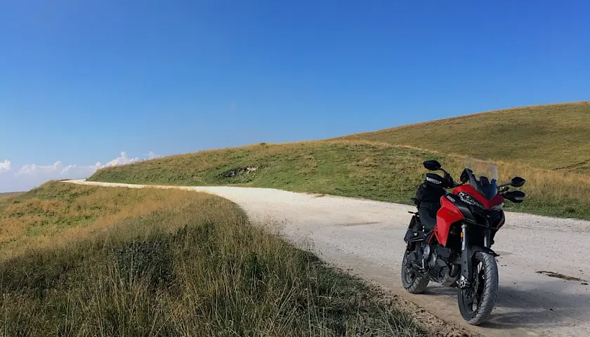 Ducati rossa ferma su strada sterrata, con un passo branchetto sullo sfondo e cielo azzurro.