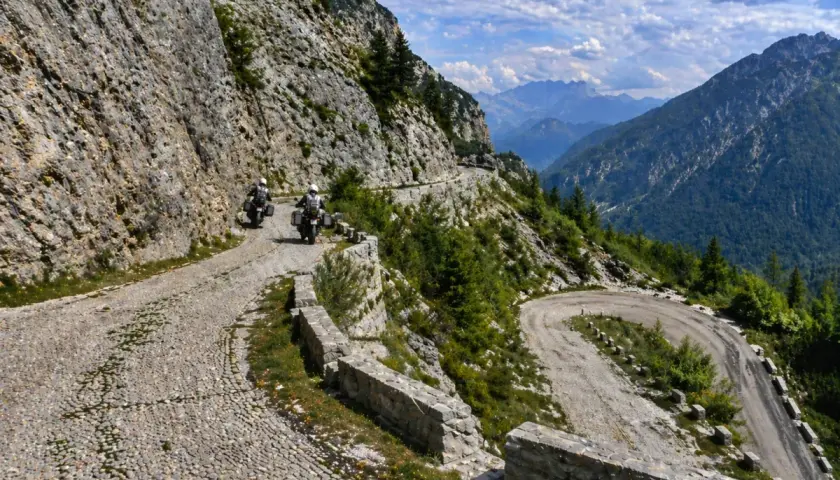 Due motociclisti affrontano tornanti mozzafiato sul passo del Tarvisio, tra rocce imponenti e valli verdi.