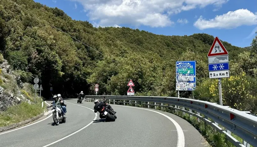 Motociclisti in curva sul passo del Bracco, tra il verde della Liguria e un cielo azzurro con nuvole bianche.