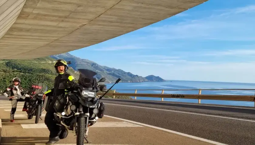 Motociclisti in sosta panoramica, con moto pronte per esplorare la costa tra Alghero e Oristano. Mare azzurro e montagne sullo sfondo.