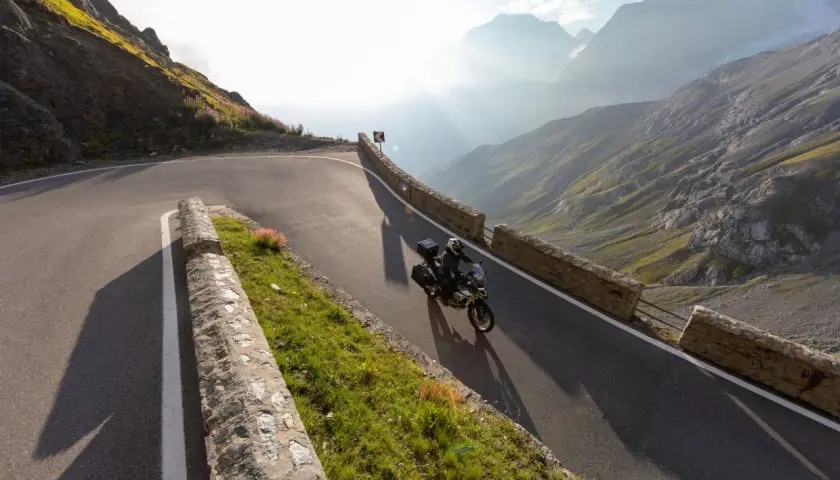 Motociclista affronta tornanti mozzafiato sul Passo Giovo, con montagne maestose e raggi di sole che illuminano il paesaggio alpino.