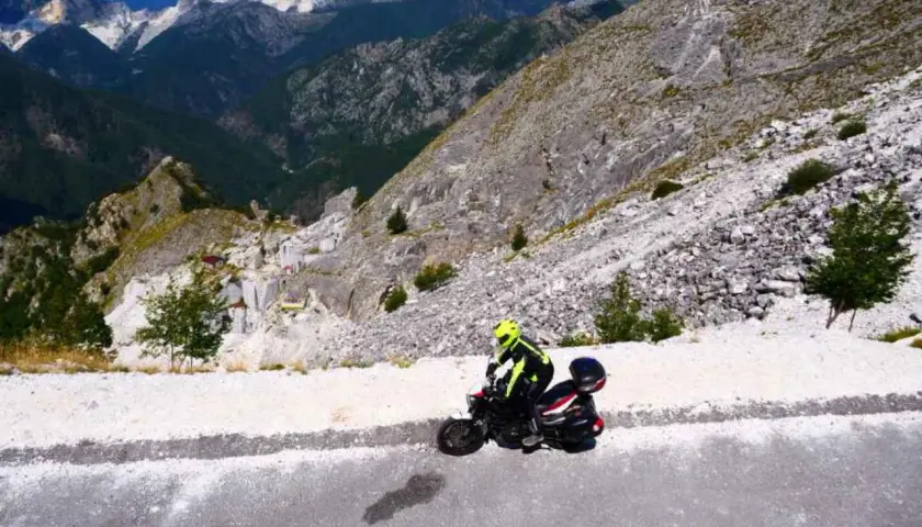 Un motociclista con casco giallo affronta il passo del vestito in moto, tra montagne rocciose e cave di marmo.