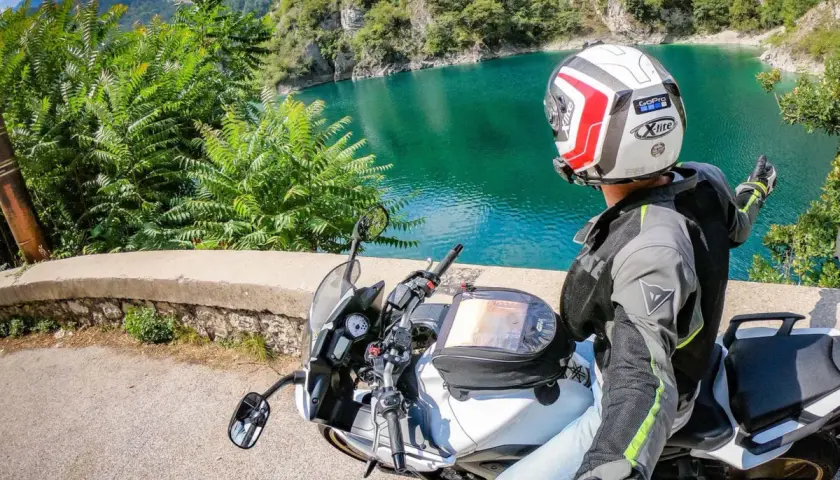 Weekend in moto nel Nord Italia: un motociclista ammira un lago alpino turchese, con montagne verdi sullo sfondo.