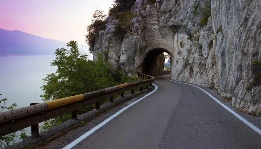 Strada della forra in moto, con un tunnel scavato nella roccia e il lago sullo sfondo al tramonto.