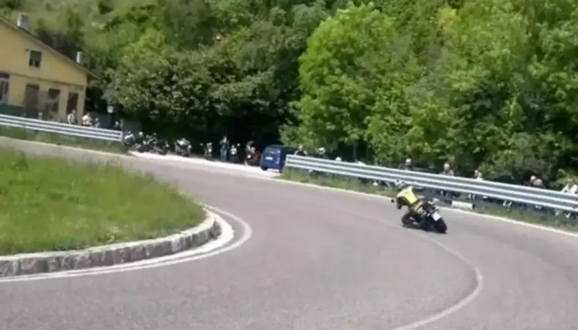 Un motociclista in giallo affronta una curva su una **strada del costo** panoramica, con spettatori e moto parcheggiate a lato.