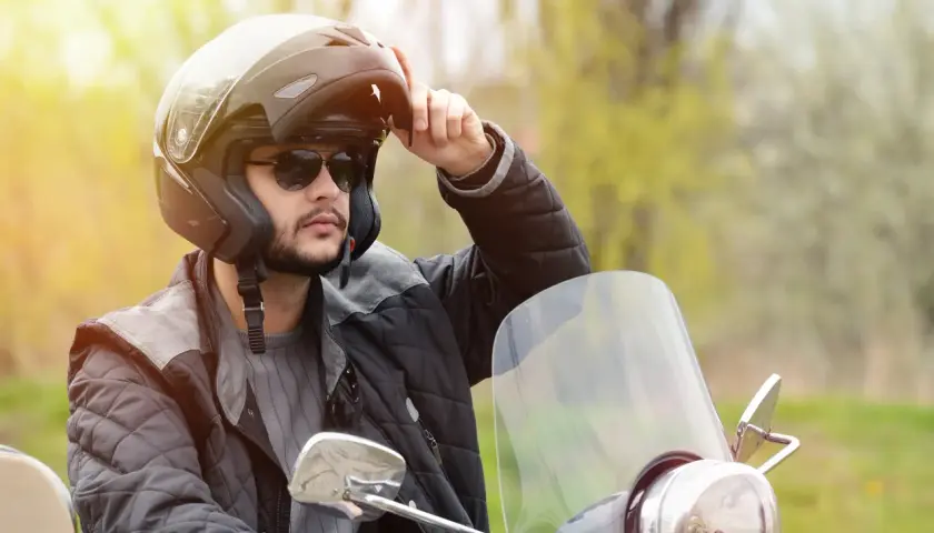 Uomo con casco da moto, occhiali da sole e giacca imbottita, che regola le parti del casco.