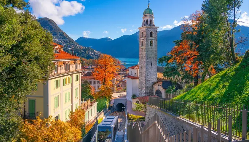 Un trenino scende tra case colorate e un campanile, con il Lago Maggiore sullo sfondo. Un assaggio delle **strade panoramiche Lago Maggiore** in autunno.