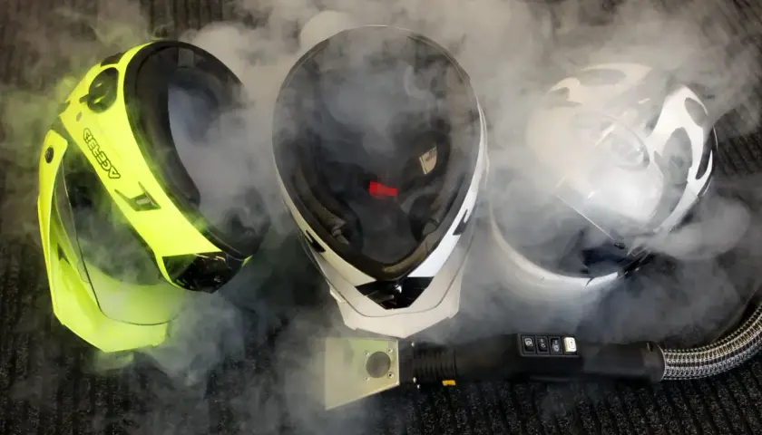 Tre caschi, uno giallo fluo, uno bianco e nero e uno bianco, circondati da vapore. Un pulitore a vapore è posizionato davanti, suggerendo che il casco non deve essere pulito con solventi.