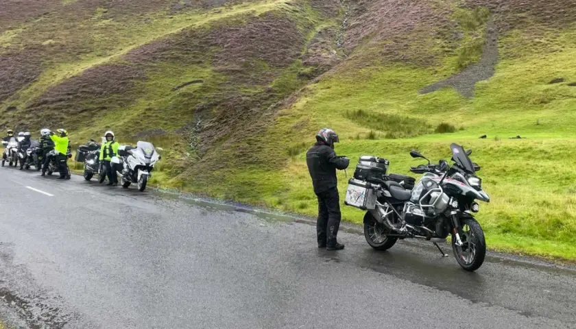 Motociclisti in sosta sul passo della Contessa, con moto cariche e un paesaggio collinare verde e viola sullo sfondo.