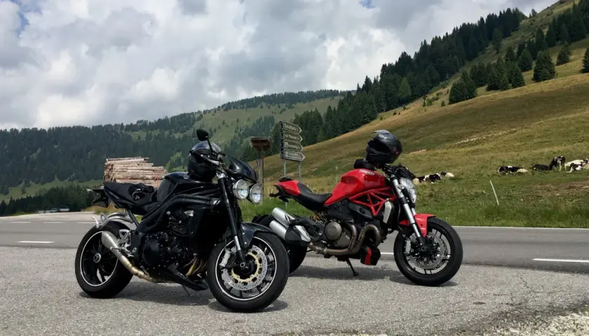 Due moto, una nera e una rossa, parcheggiate sul ciglio della strada al passo Castrin. Mucche al pascolo sullo sfondo di un prato verde e alberi.