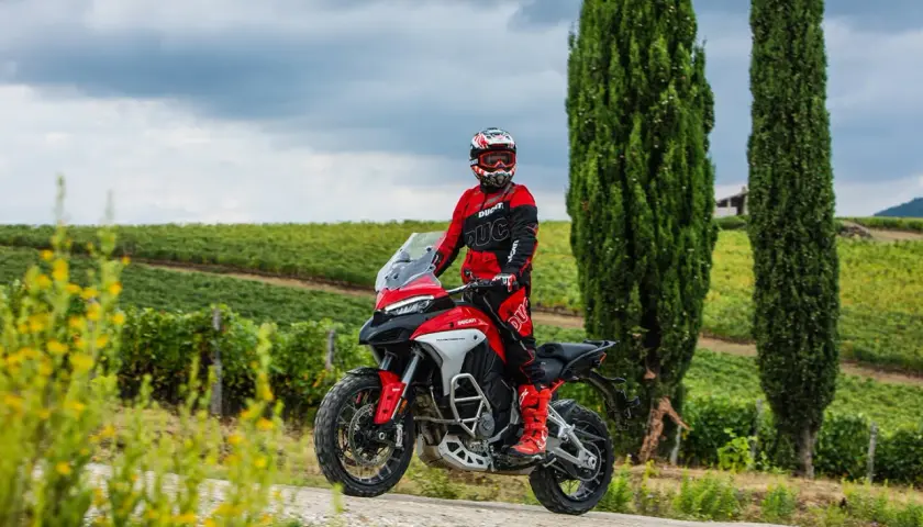 Un motociclista in tuta rossa sfreccia tra i cipressi e i vigneti del Val d'Orcia in moto.