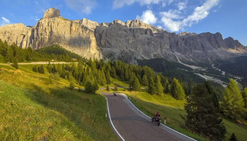 Strada tortuosa tra prati verdi e boschi di conifere, con imponenti vette dolomitiche sullo sfondo. Due motociclisti godono del panorama mozzafiato, un'avventura indimenticabile tra le Dolomiti in moto.