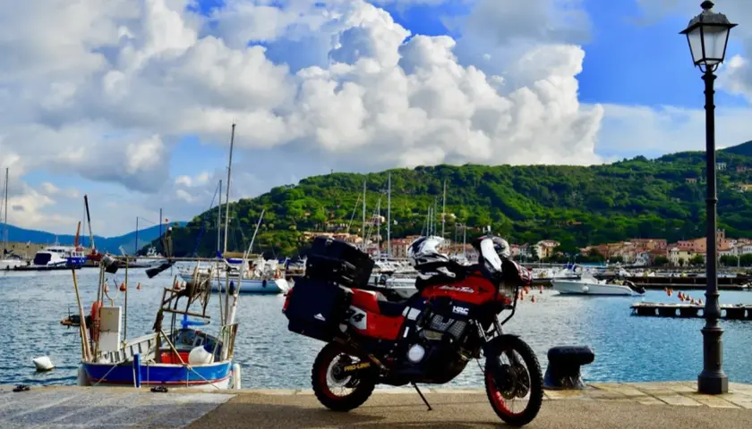 Moto pronta per l'avventura: Isola d'Elba in moto 3 giorni. Porto con barche e collina verde sullo sfondo.