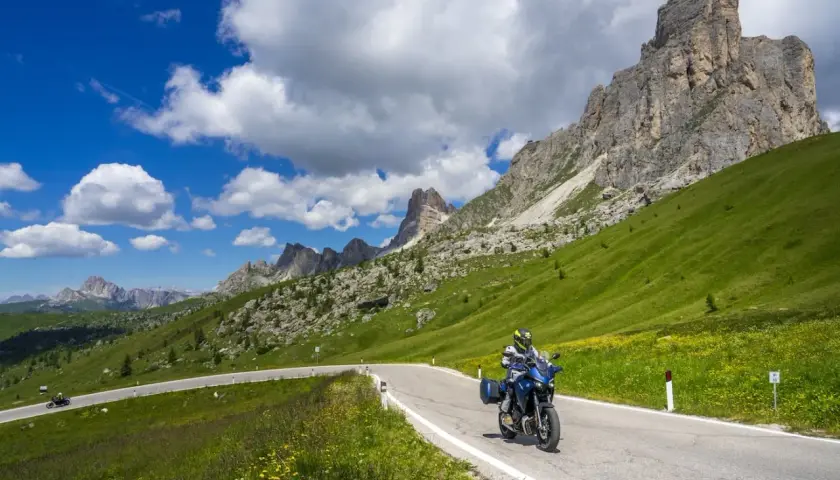 Motociclista percorre una strada panoramica tra i laghi del Veneto, con montagne imponenti e cielo azzurro.