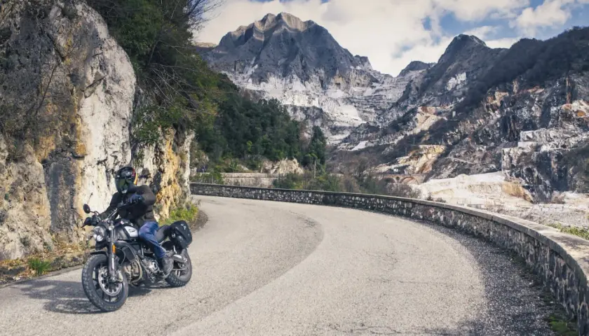 Motociclista su strada panoramica toscana, con montagne di marmo sullo sfondo.
