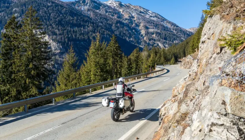 Un motociclista percorre una strada tortuosa in Valtellina, circondato da imponenti montagne e fitti boschi di conifere.