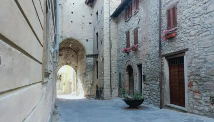 Un vicolo suggestivo a Varzi, con un arco in pietra che conduce a una strada illuminata. Case antiche e fiori colorati invitano a scoprire cosa vedere.