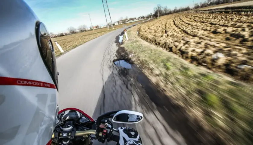 In sella a una moto, con il casco bianco e rosso, si percorre una strada di campagna. Il campo arato a lato e il cielo azzurro evocano il linguaggio motociclisti italiano.