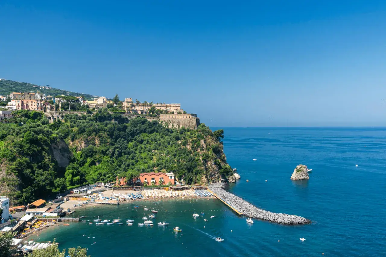 Vico Equense cosa vedere: una baia con spiaggia attrezzata, un molo di rocce e un borgo arroccato sulla scogliera.