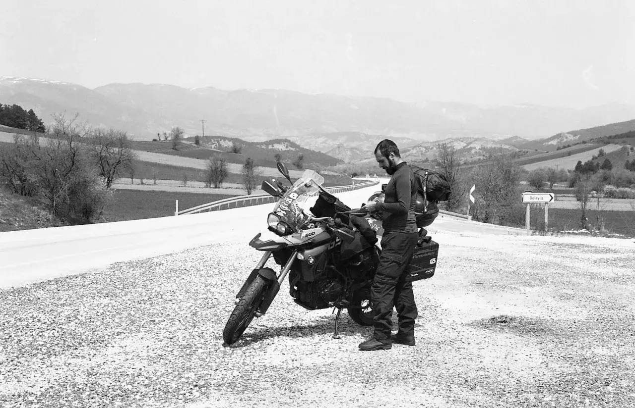 Motociclista con zaino e attrezzatura da viaggio si ferma accanto alla sua moto, la sella ciampigotto pronta per la prossima avventura. Paesaggio collinare sullo sfondo.