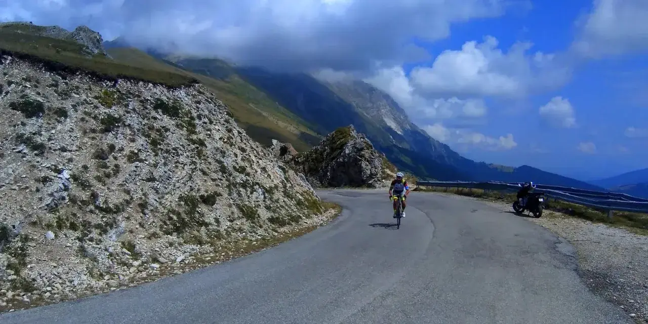 Ciclista in salita sulla strada panoramica della Forca di Presta, con montagne sullo sfondo e una moto parcheggiata.