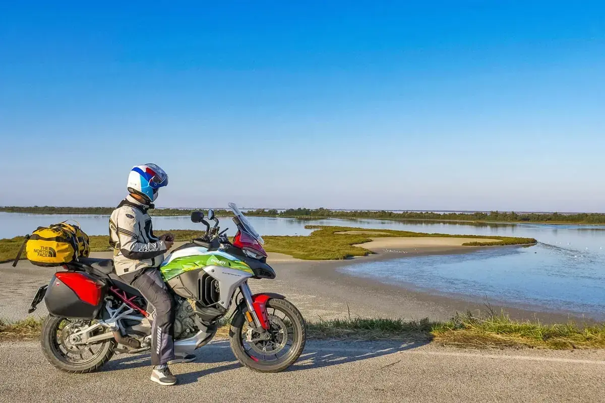 Motociclista con moto da turismo si ferma lungo la strada statale 9 Via Emilia, con una laguna sullo sfondo.