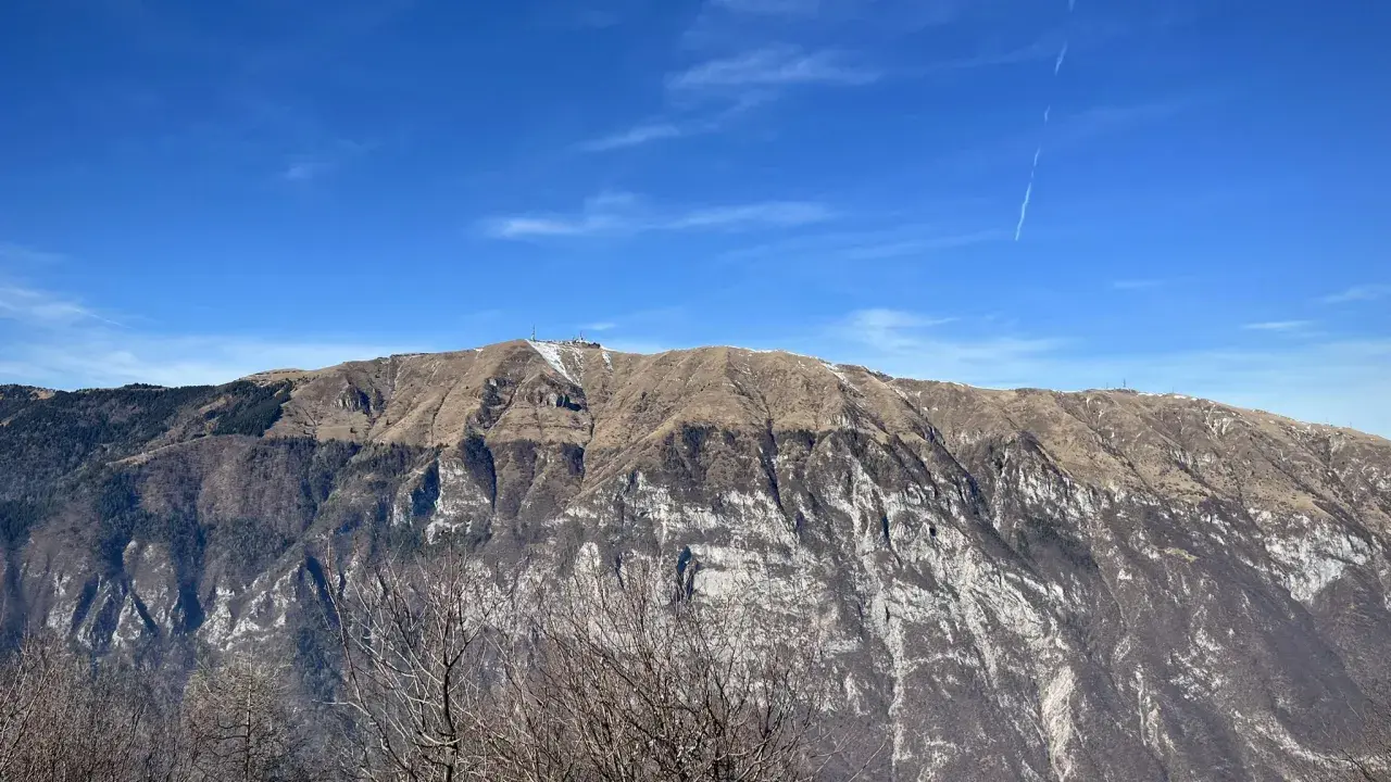 Vetta del monte Pizzoc con neve residua, pendii bruni e rocce bianche sotto un cielo azzurro con scie di aerei.