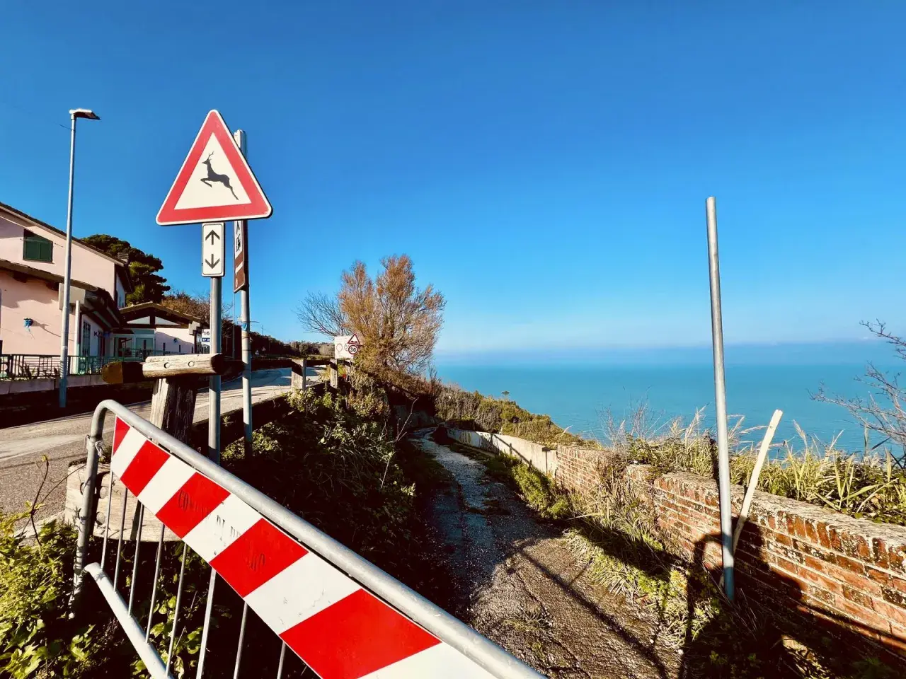 Panoramica Gabicce con segnale di pericolo cervi e mare azzurro. Strada sterrata con muretto in mattoni.