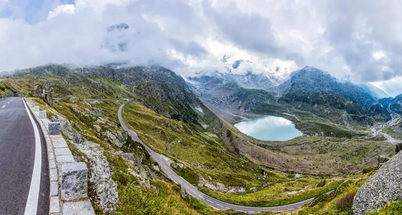 Strada tortuosa sul passo della Sambuca, con lago alpino azzurro e montagne innevate sullo sfondo.
